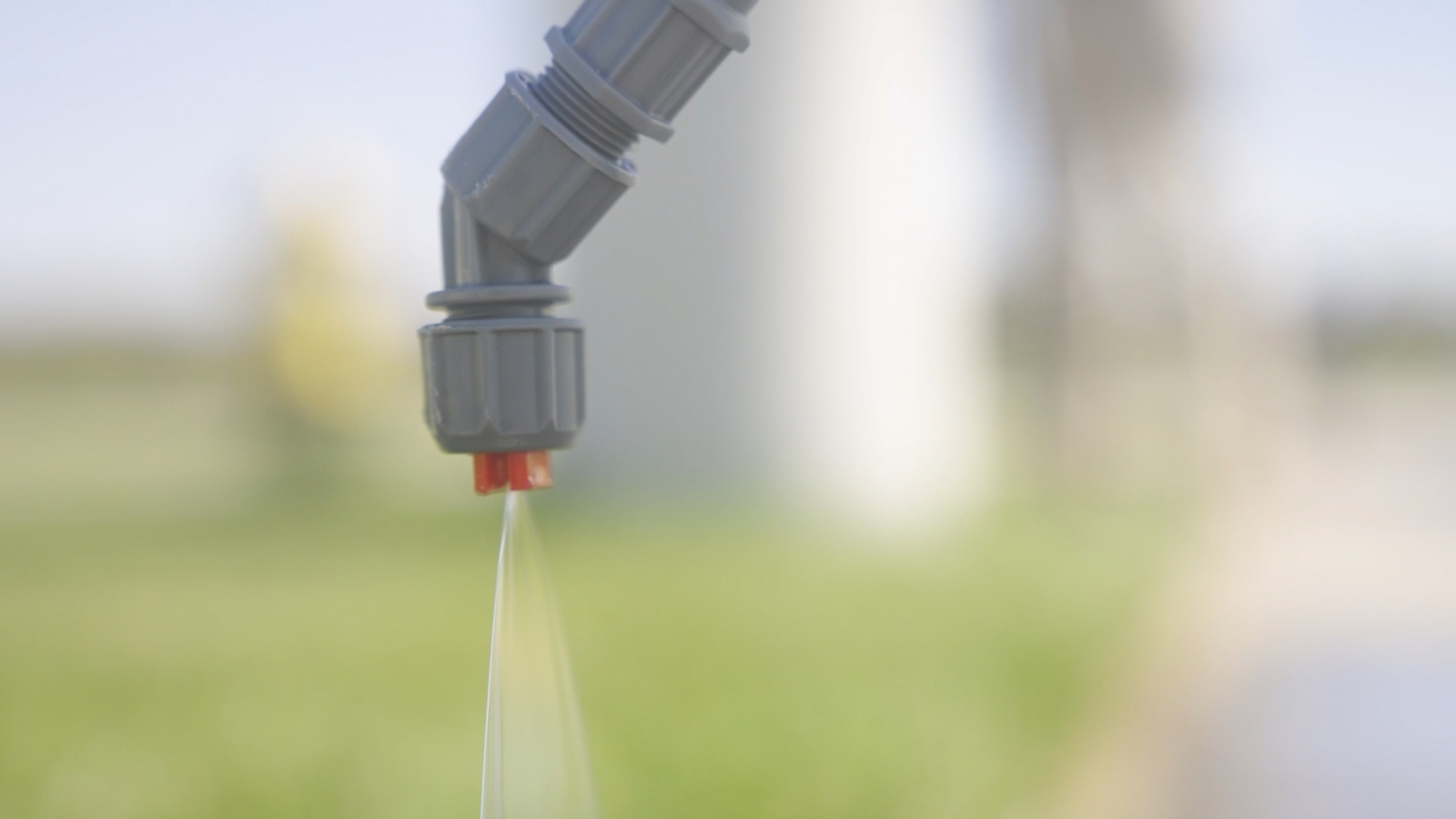 closeup image of sprayer nozzle with liquid spraying out, set against a background of blurred grass and farm silo.